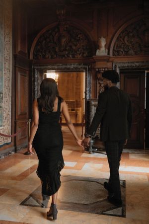 Couple in elegant black attire walking through an ornate hallway with decorative arches and marble floors.
