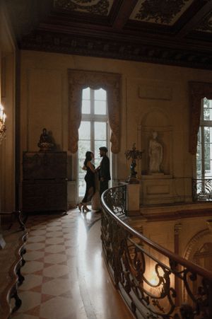 A couple dances in silhouette against a bright window in an elegant historic ballroom with ornate railing and marble floor.