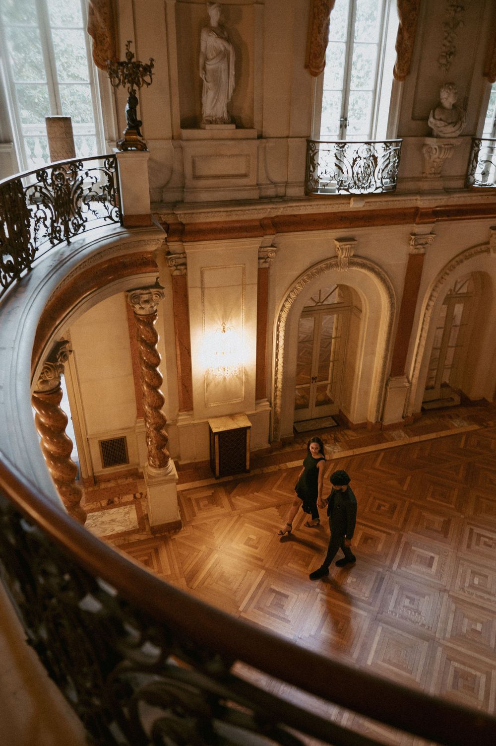 Looking down a curved marble staircase in an elegant palace with decorative railings and classical statues.