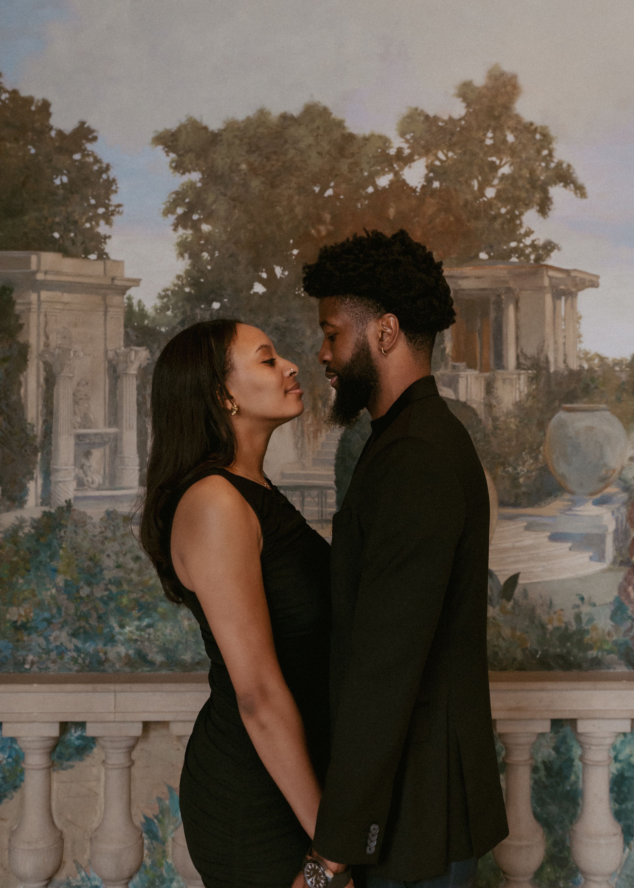 A romantic couple in all black outfits share an intimate moment against an ornate garden backdrop with classical architecture.