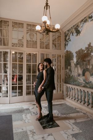 A couple in black outfits stands in an elegant room with vintage wallpaper, ornate windows and a chandelier overhead.