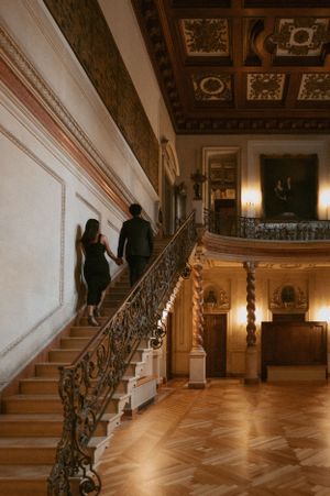 A couple ascends an ornate marble staircase in a luxurious historic mansion with detailed wood coffered ceilings.