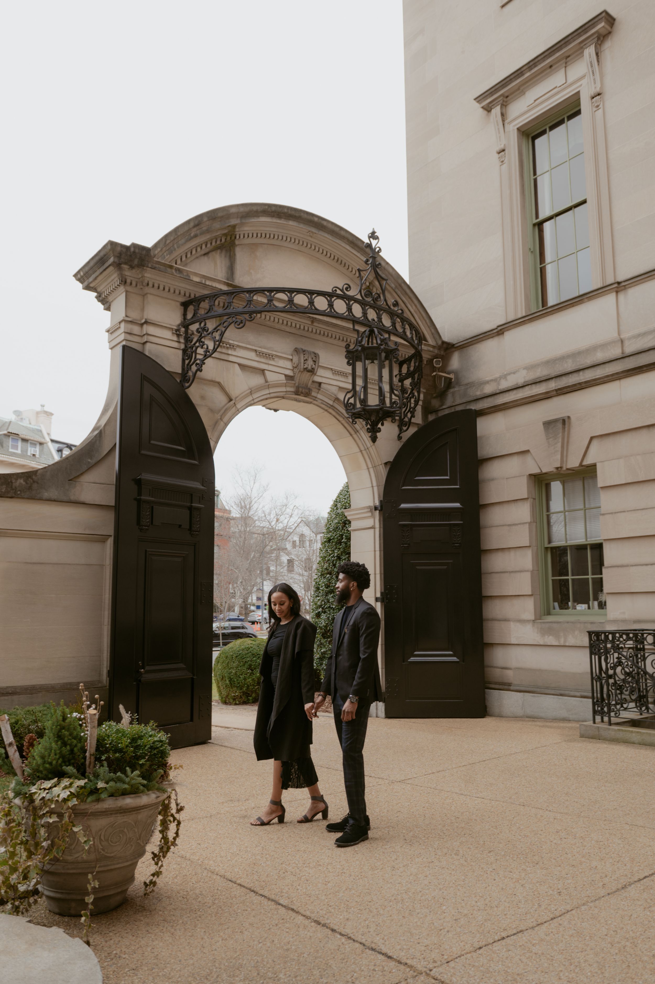 A couple stands beneath an elegant arched entrance with ornate architectural details of a historic building.