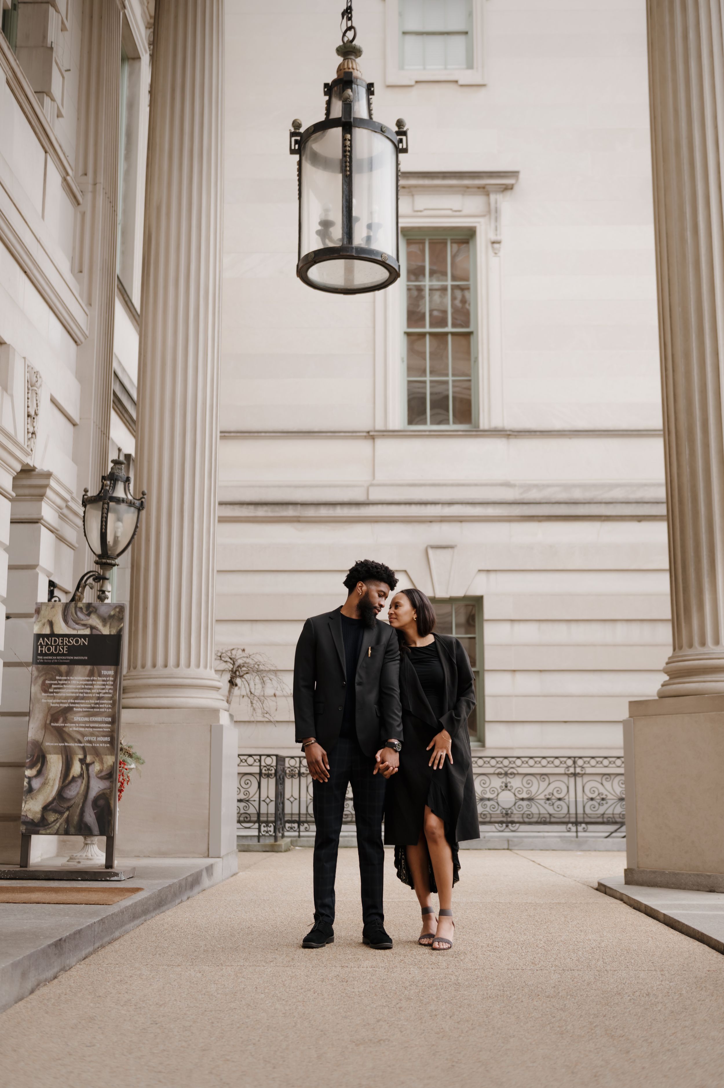 Couple in black coats standing under ornate classical columns and hanging lanterns of historic building.