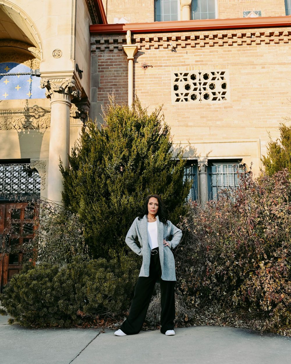 Outdoor fashion photo by beige stone building with ornate details and decorative shrubs.
