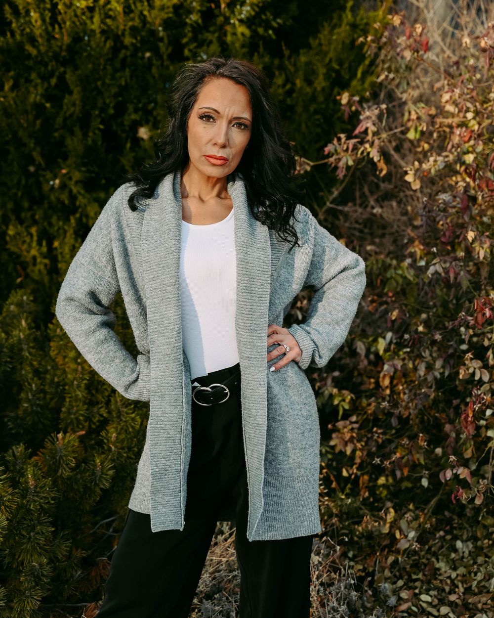 A casually styled woman poses outdoors in a grey cardigan and white top against an autumn background.