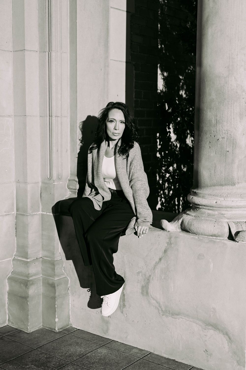 Black and white portrait of someone sitting casually on stone steps by columns wearing a light jacket and dark pants.