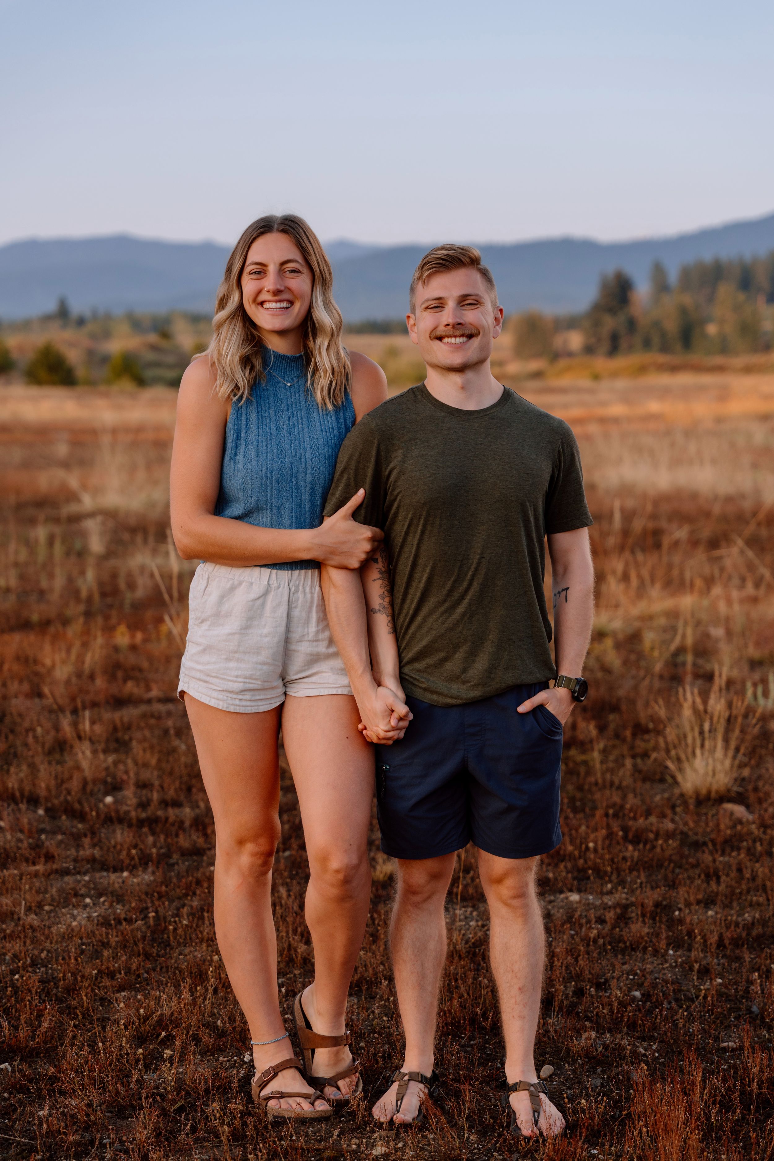 Two photographers stand together against white wall holding professional camera equipment and smiling at camera.