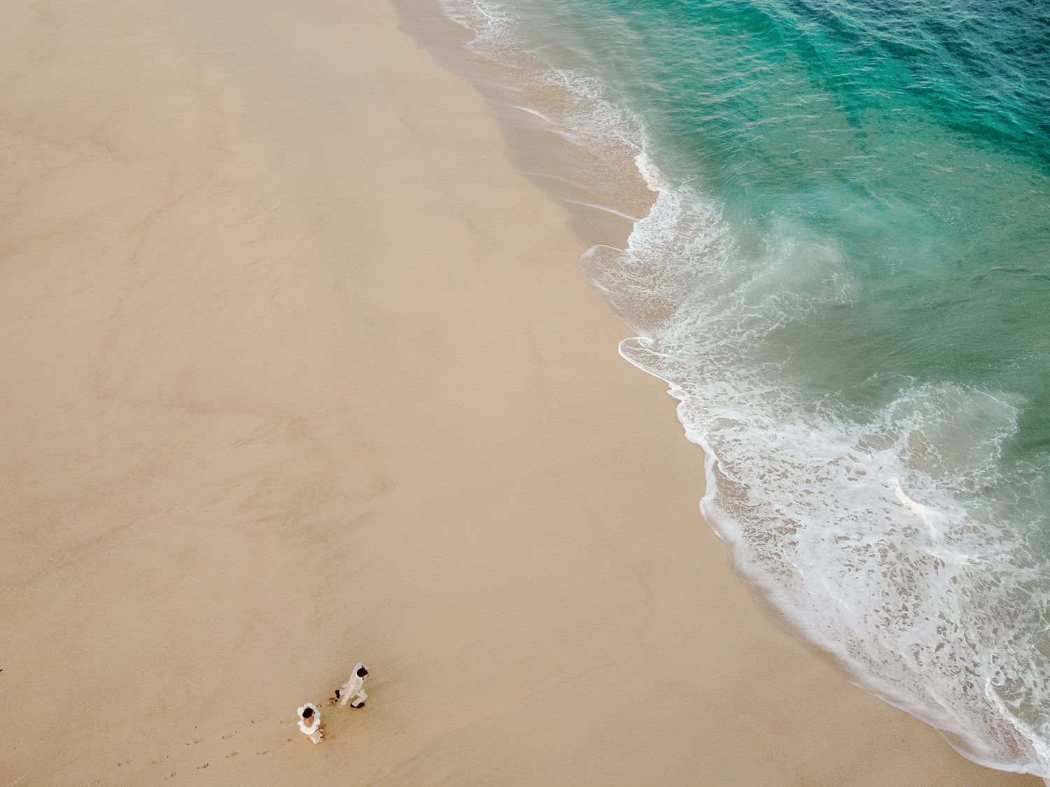 Aerial view of two beachgoers walking along pristine sandy shoreline with turquoise ocean waves rolling in.