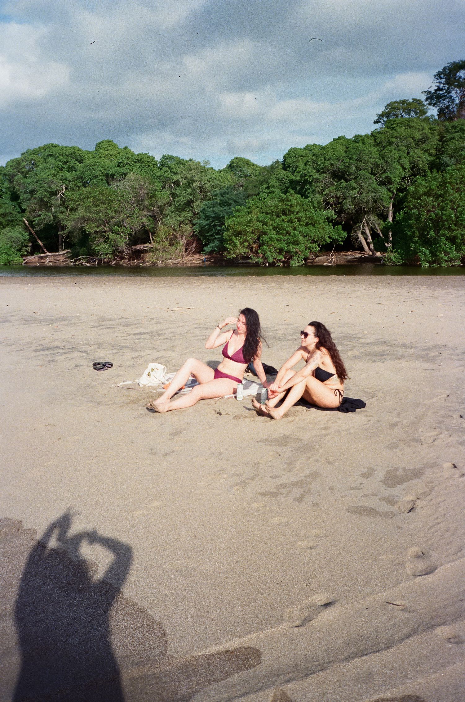 Two beachgoers relax on a sandy shore near lush green trees and murky brown water on an overcast day.