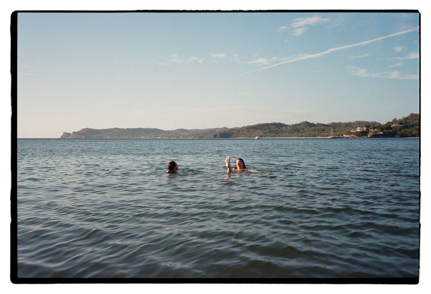 People swimming in calm blue waters of a coastal bay surrounded by rugged terrain on a sunny summer day.