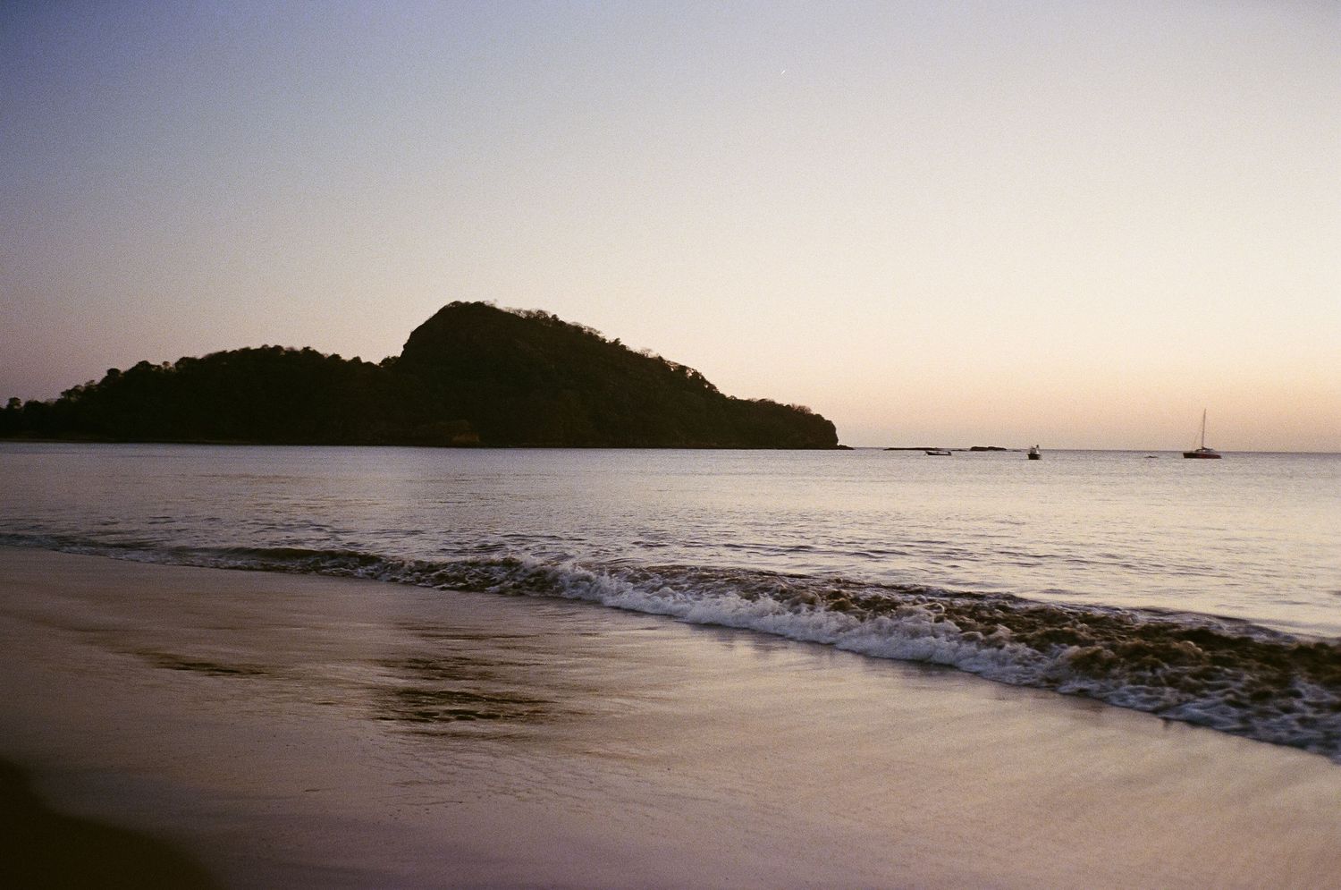 Serene beach landscape at sunset with silhouetted island and gentle waves lapping at sandy shore.