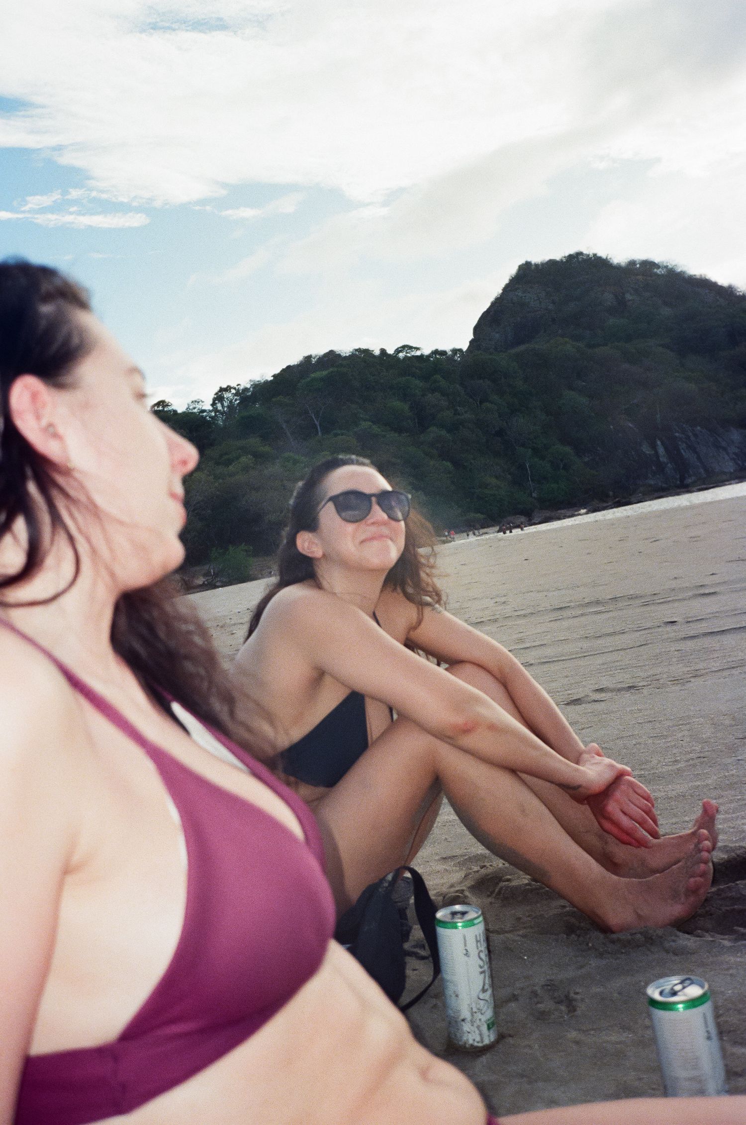 Friends relaxing and laughing together on a scenic beach with beverages and mountains in the background.