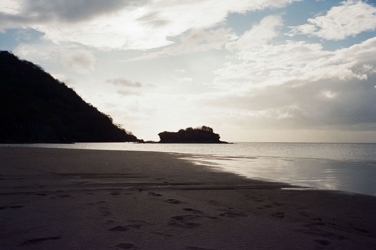 Dramatic cloudy sky over deserted beach with dark mountain silhouette stretching into calm ocean waters.