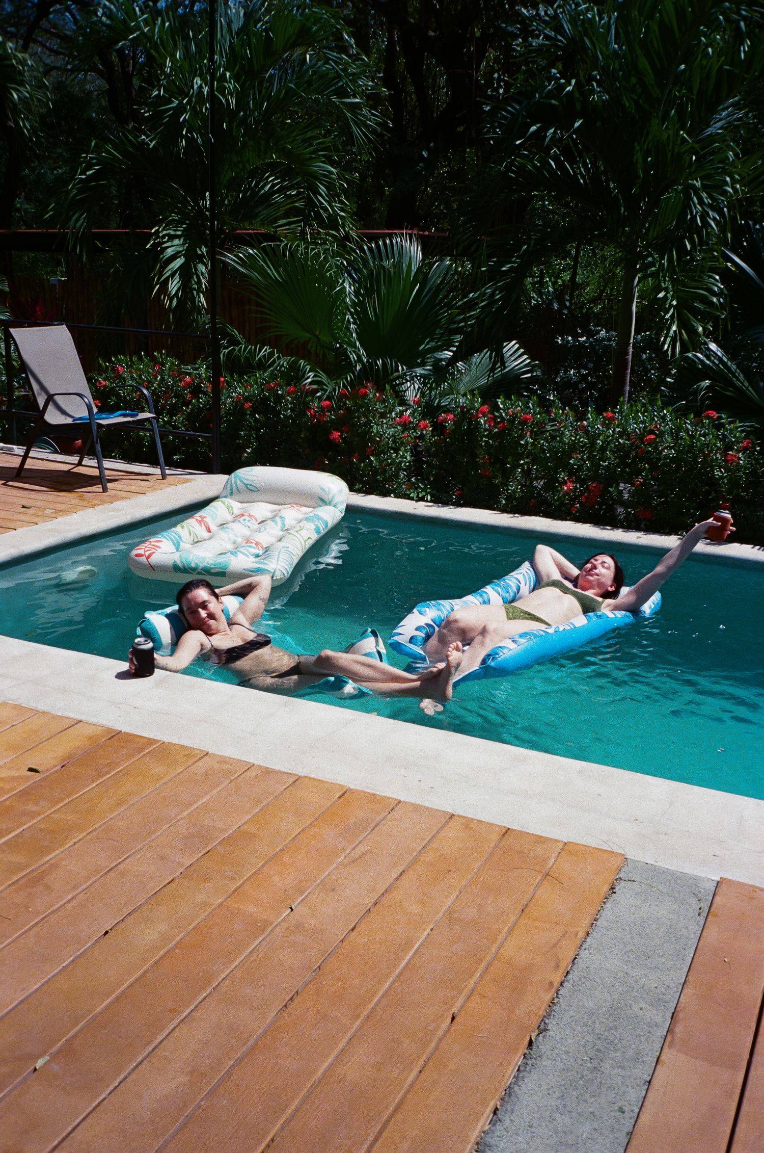 People relaxing on colorful pool floats in a backyard swimming pool surrounded by tropical plants and wooden decking.