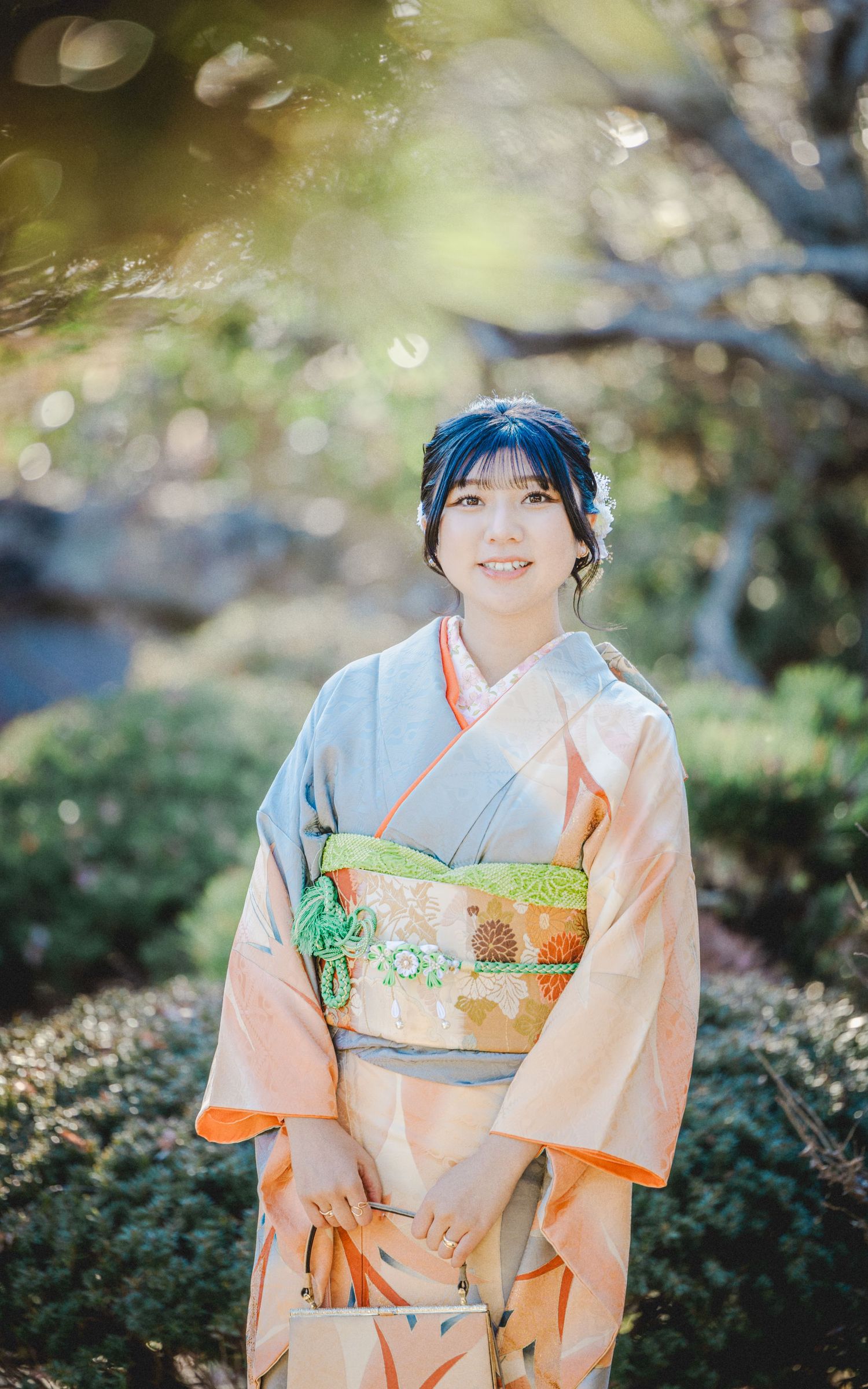 A person wearing a peach-colored kimono with floral obi stands in a sunlit garden setting.