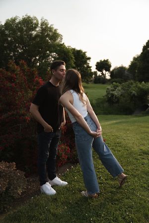 Two people interact playfully on a grassy hill with red flowering bushes nearby.