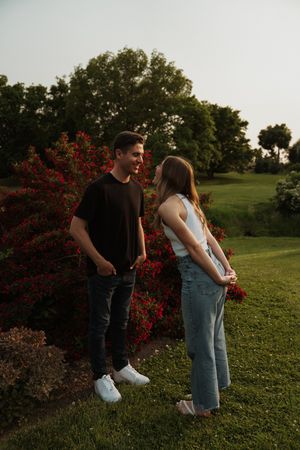 Two people share a moment together on a grassy slope with red flowering bushes.