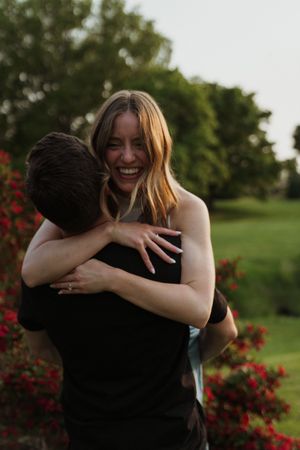 A joyful embrace is shared between two people near flowering red bushes in a garden.