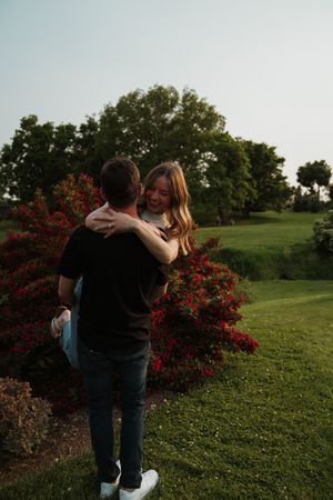 Romantic couple embraces in a lush green field with blooming red flowers at sunset.