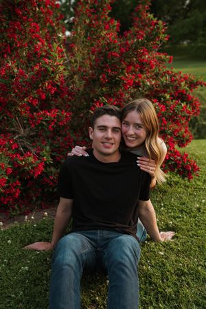 A couple sits together on grass near vibrant red flowering bushes in a garden setting.
