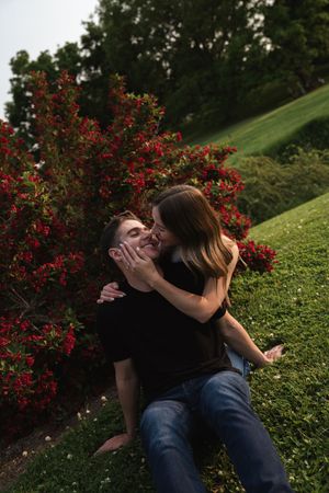 Two people share a playful moment while sitting by red flowering bushes.