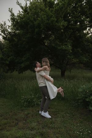 Couple embracing and lifting each other playfully in a green outdoor setting.