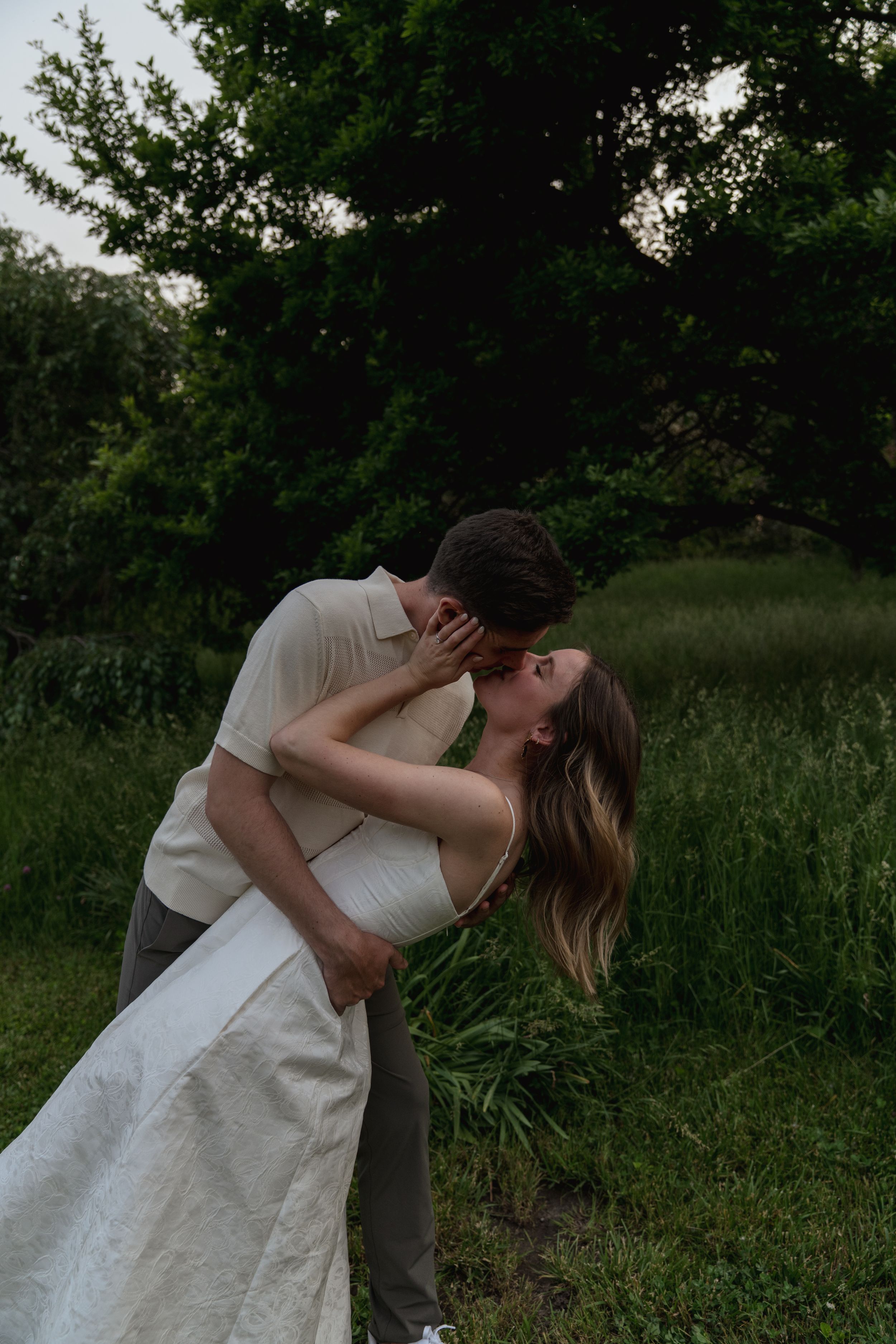 A romantic couple shares a tender dip kiss in a lush green outdoor setting.