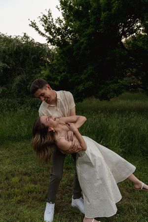 A romantic couple in white and gray outfits dancing and dipping in a grassy outdoor setting at dusk.