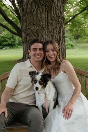 A couple sits on a wooden bench with their Australian Shepherd dog against a large tree trunk.