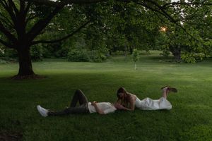 A couple relaxes on their backs in the grass under a large tree in a peaceful park.