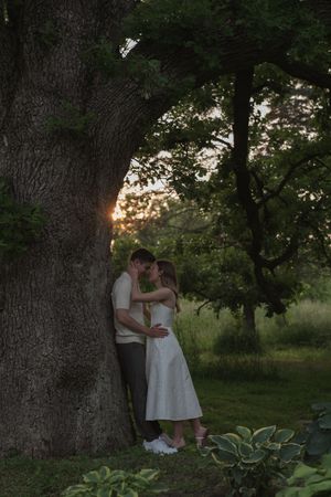 A couple shares an intimate moment beside a large tree trunk at dusk.