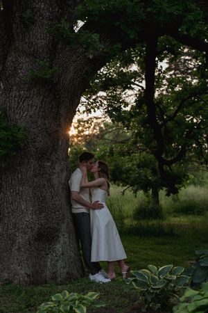 Two people embrace near a majestic tree trunk with hostas growing beneath.