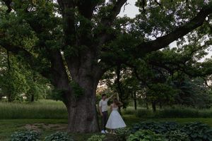 A couple stands beneath a large tree at dusk in a romantic garden setting.