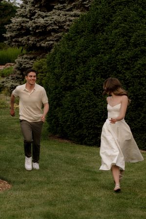 Two people in light colored clothing run playfully through a green lawn.