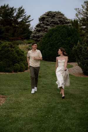 A couple runs together on a manicured lawn surrounded by evergreen trees.