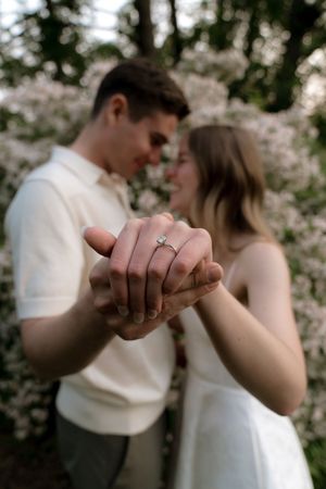 Close-up of hands showing engagement rings against a blurred floral background.