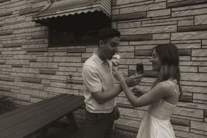 Two people share ice cream cones while sitting outdoors near a stone facade.