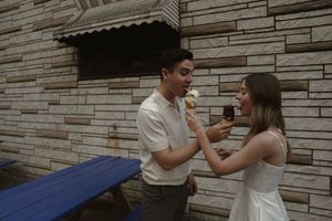 A couple in white clothing shares dessert while sitting on a bench outdoors.
