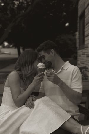 Two people in white clothing share ice cream cones while sitting together outdoors.