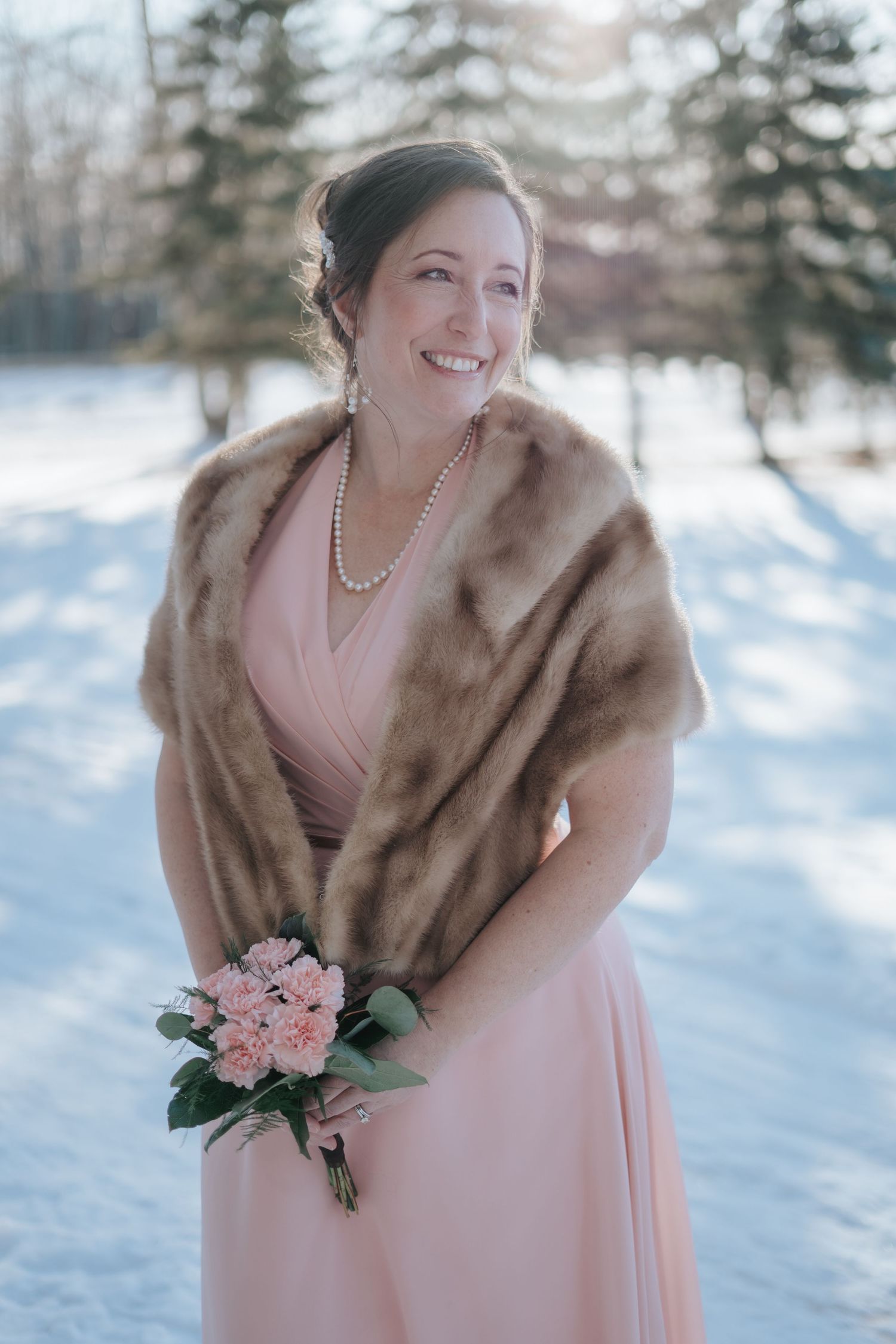 Bride in a pink dress and fur wrap holds bouquet in snowy winter setting.