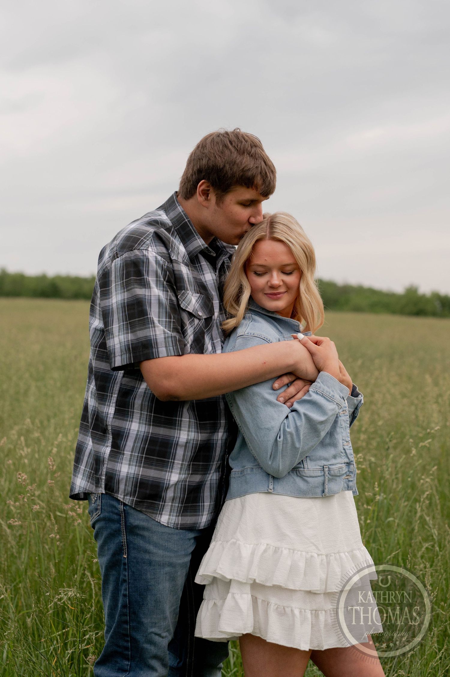A romantic couple embraces in a green meadow on an overcast day, wearing casual plaid and denim clothing.