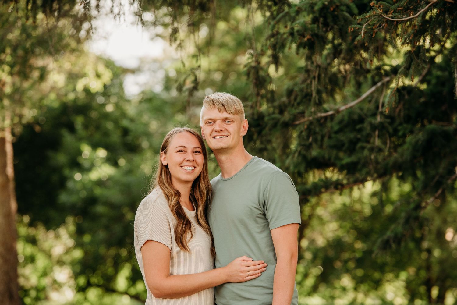 A couple embraces while standing in a sunlit wooded area on a warm summer day.