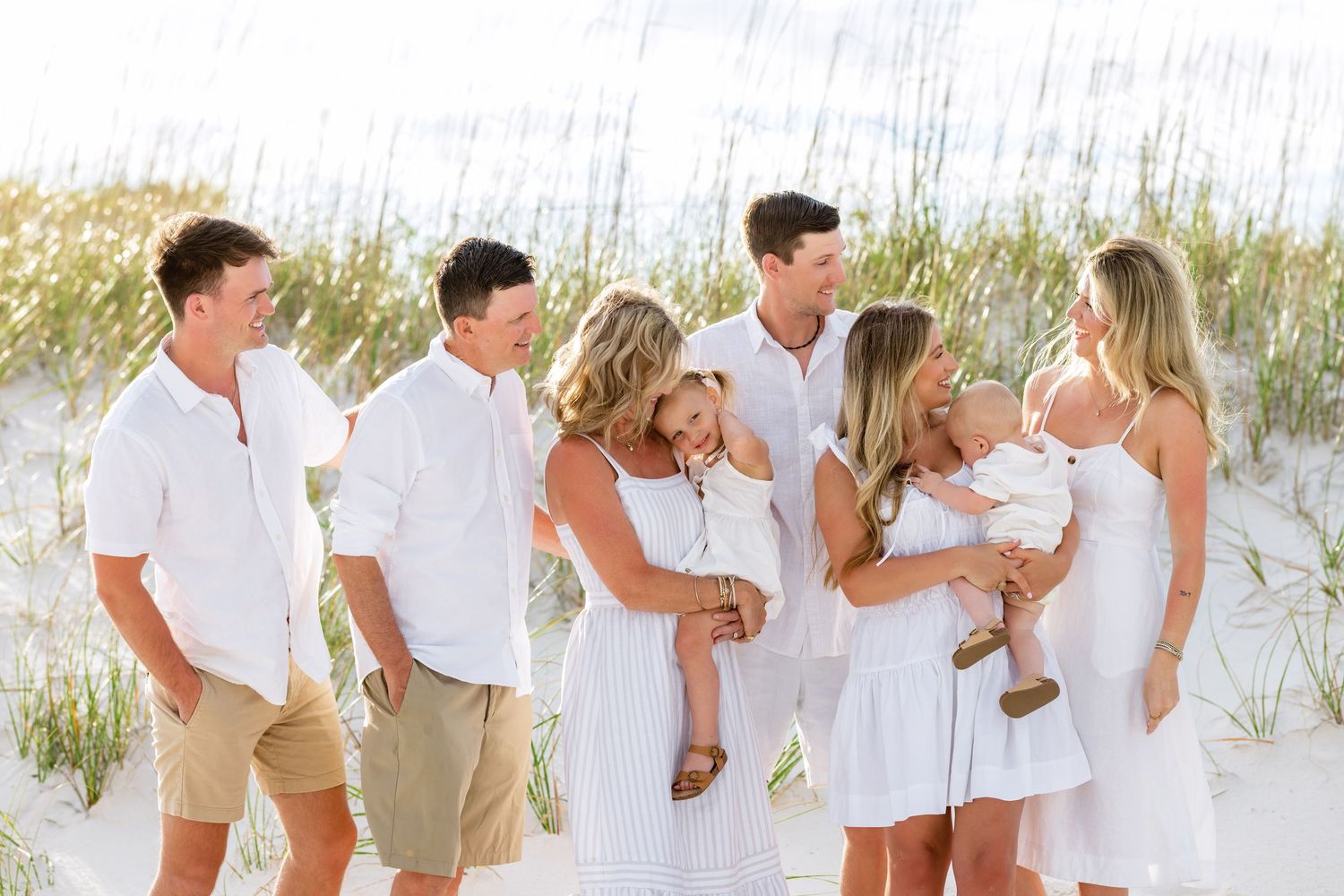 A group wearing coordinated white outfits poses together on a sandy beach with sea grass in the background.