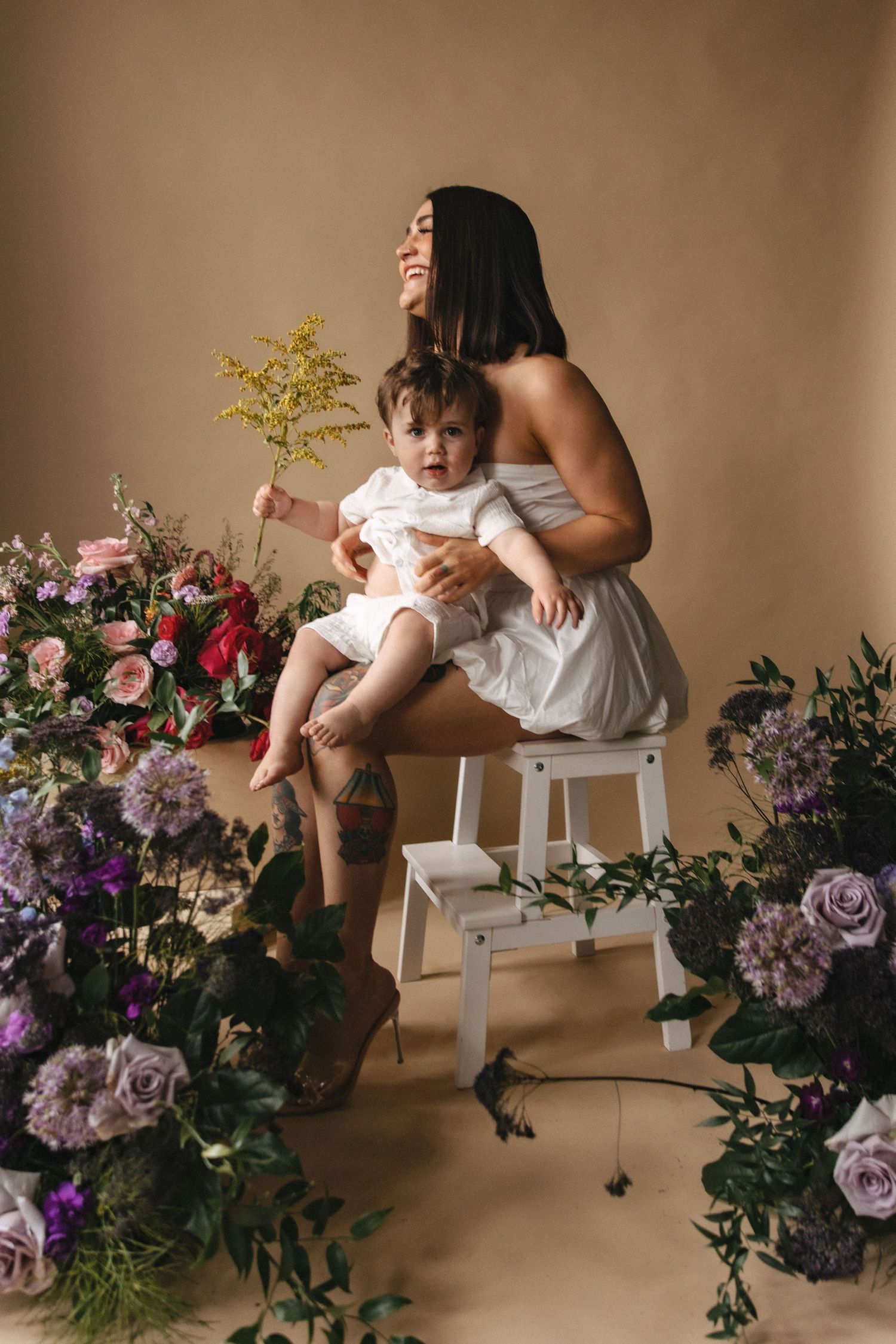 Mother and baby in white dresses sit surrounded by pink and purple flower arrangements in soft studio lighting.
