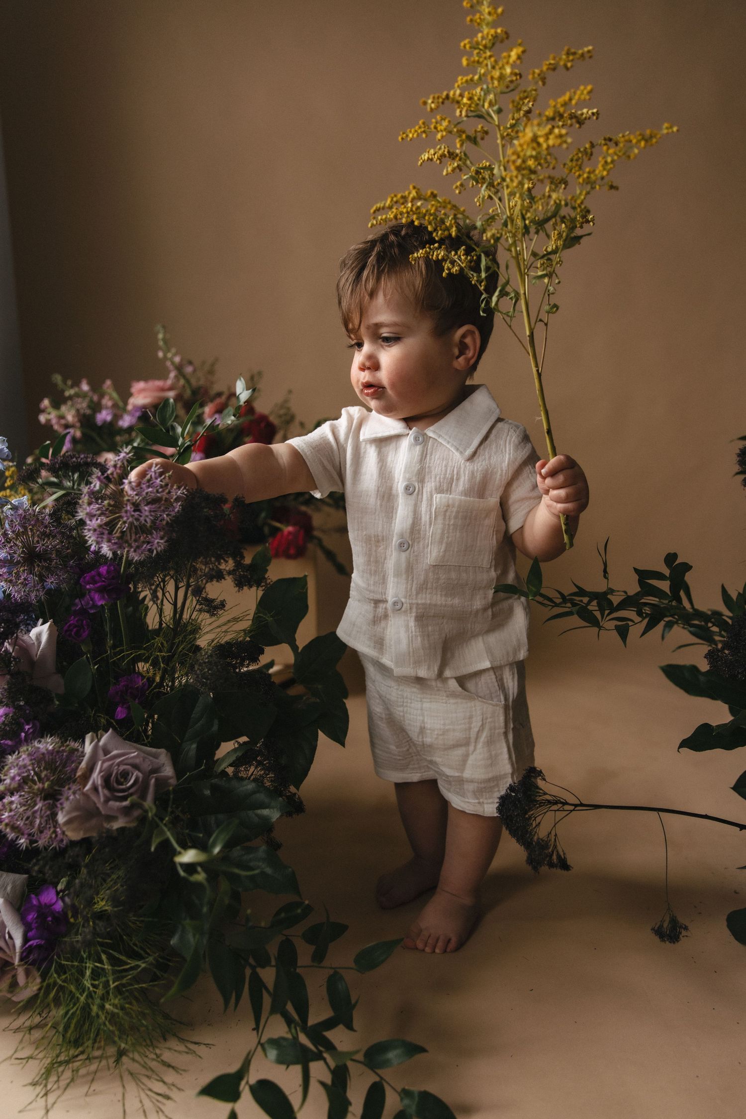 A small child in white linen clothing stands barefoot among purple and pink floral arrangements holding yellow flowers.