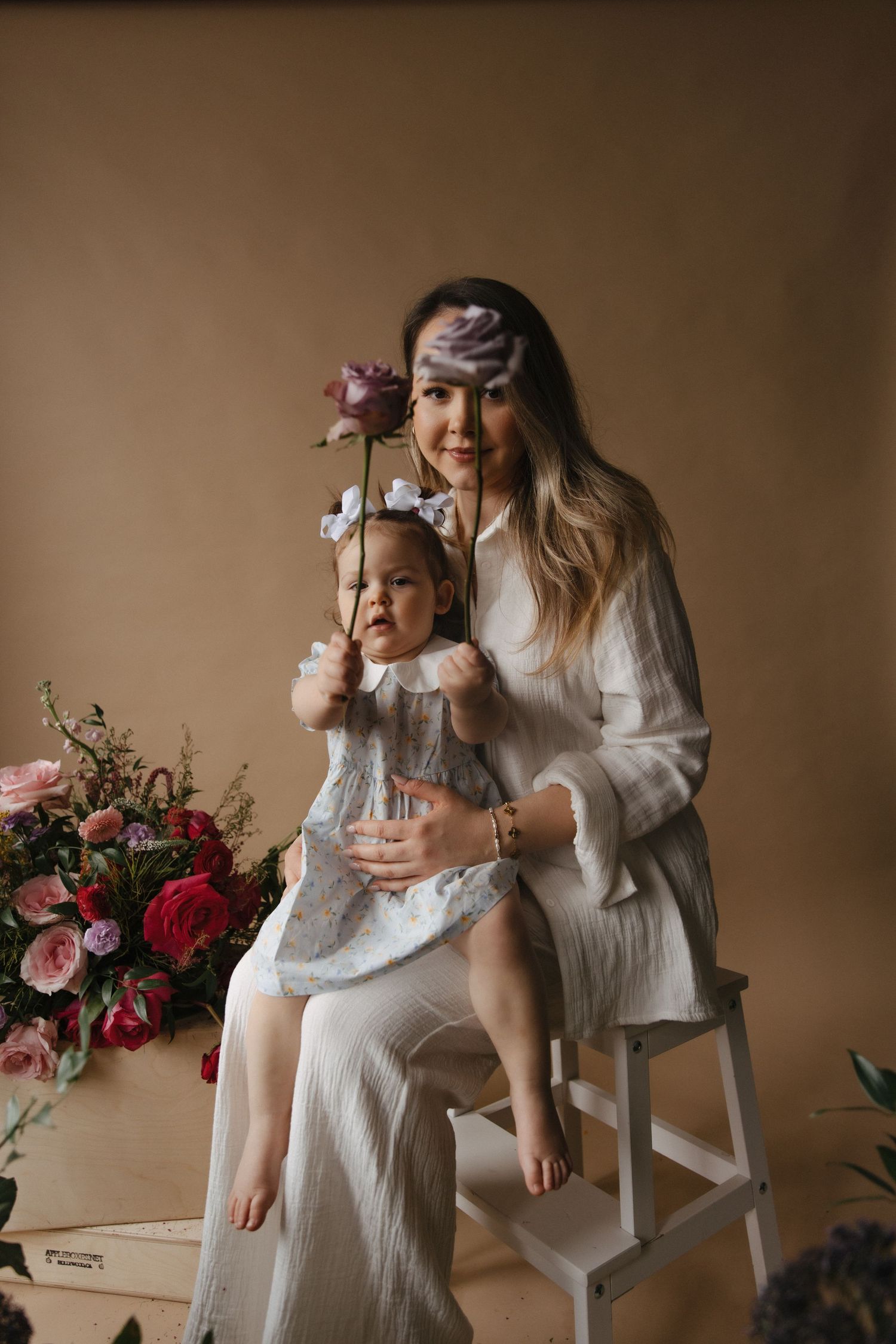 A parent and child in white dresses sit together with floral decorations in a warm-toned studio setting.
