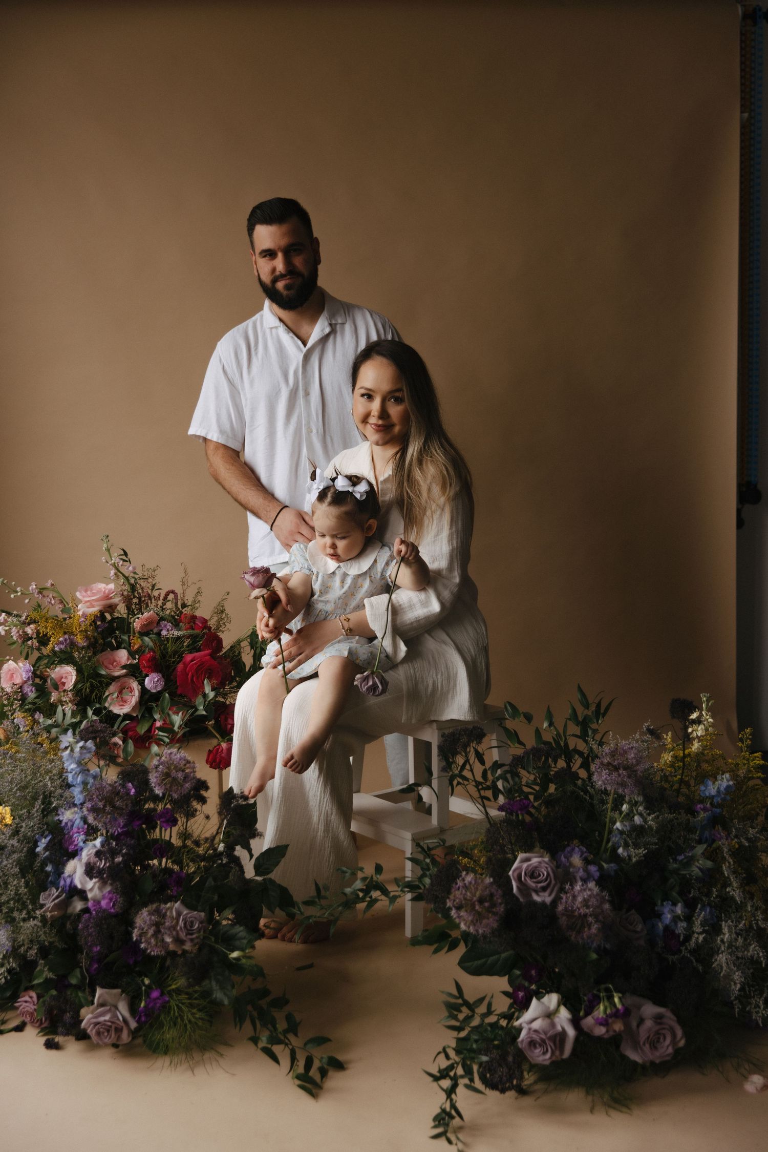 Intimate studio portrait with beautiful floral arrangements featuring roses and purple blooms against a warm beige background.