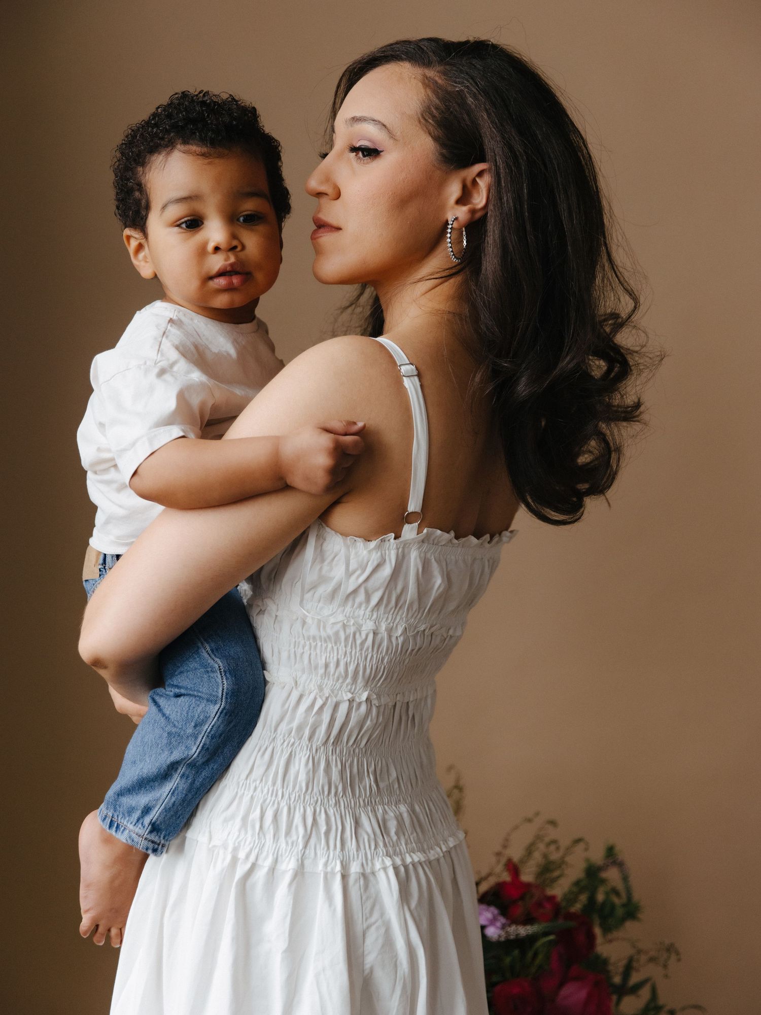 A person wearing a white smocked dress holds a young child in blue jeans against a brown studio backdrop with flowers.