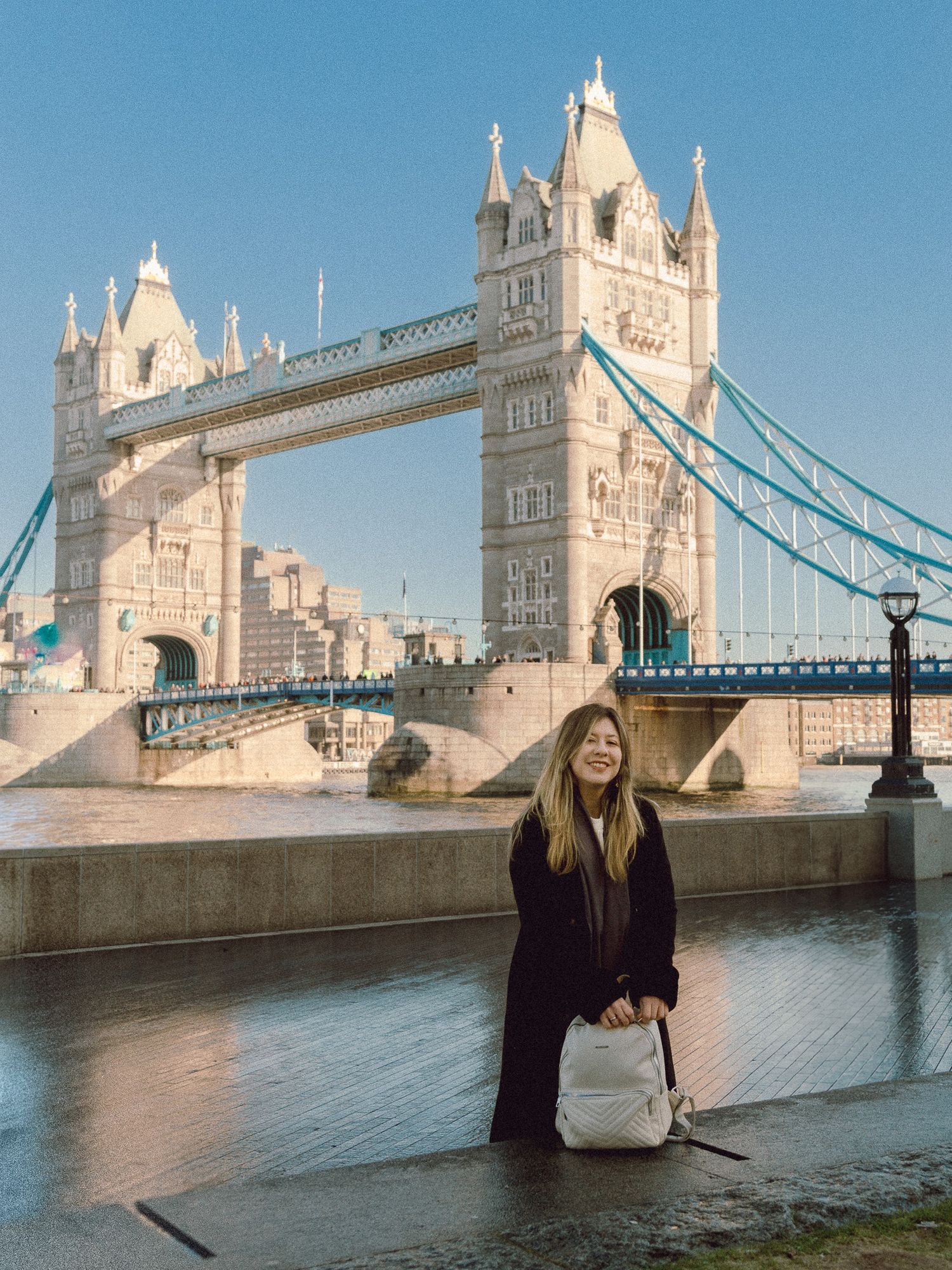 Tourist in black coat with white backpack poses near Tower Bridge on Thames riverbank.